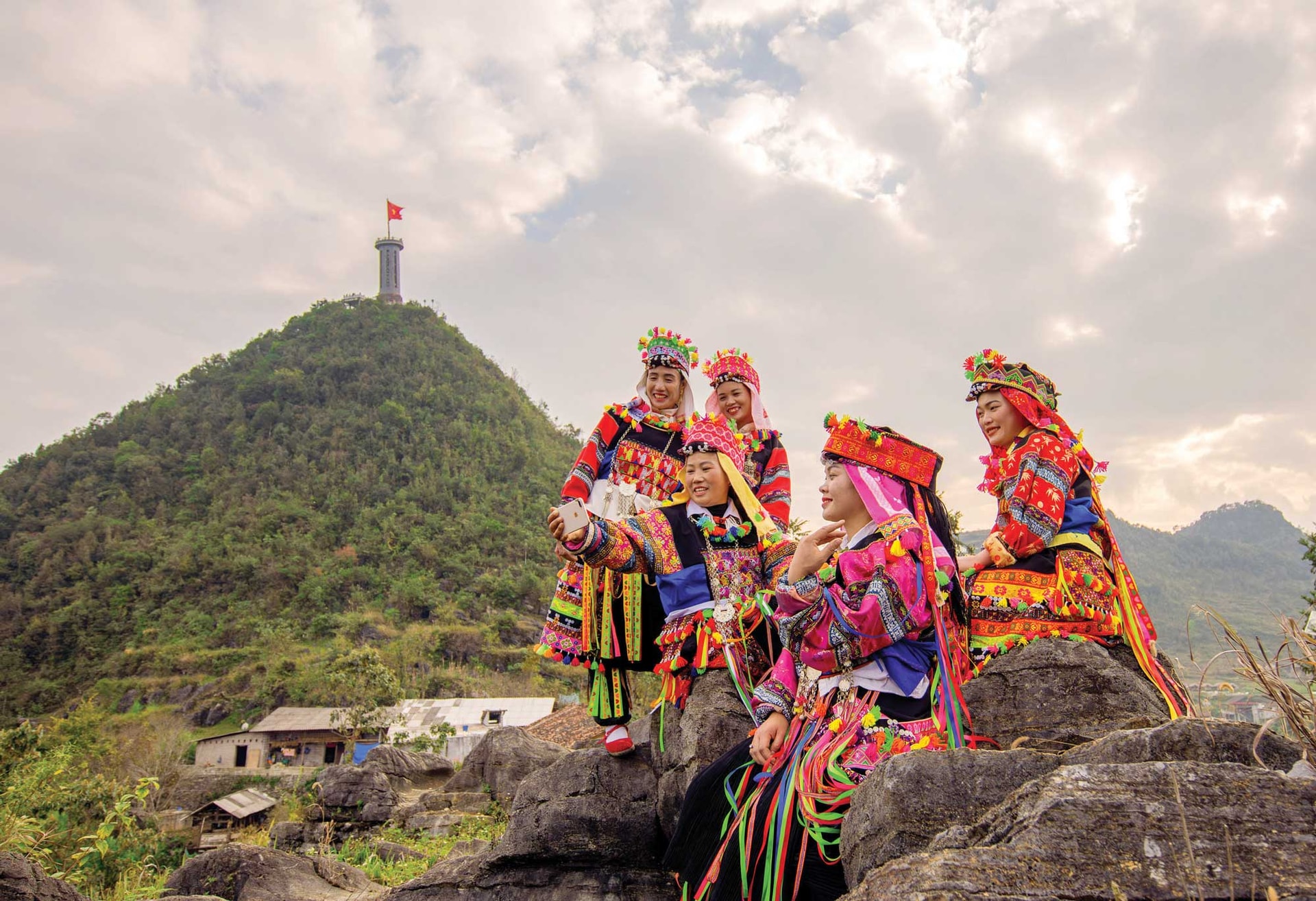 Những thiếu nữ Lô Lô trong trang phục truyền thống, phía sau là Cột cờ quốc gia Lũng Cú - điểm cực Bắc thiêng liêng, biểu tượng hào hùng của chủ quyền quốc gia_Ảnh: happy.vietnam.vn